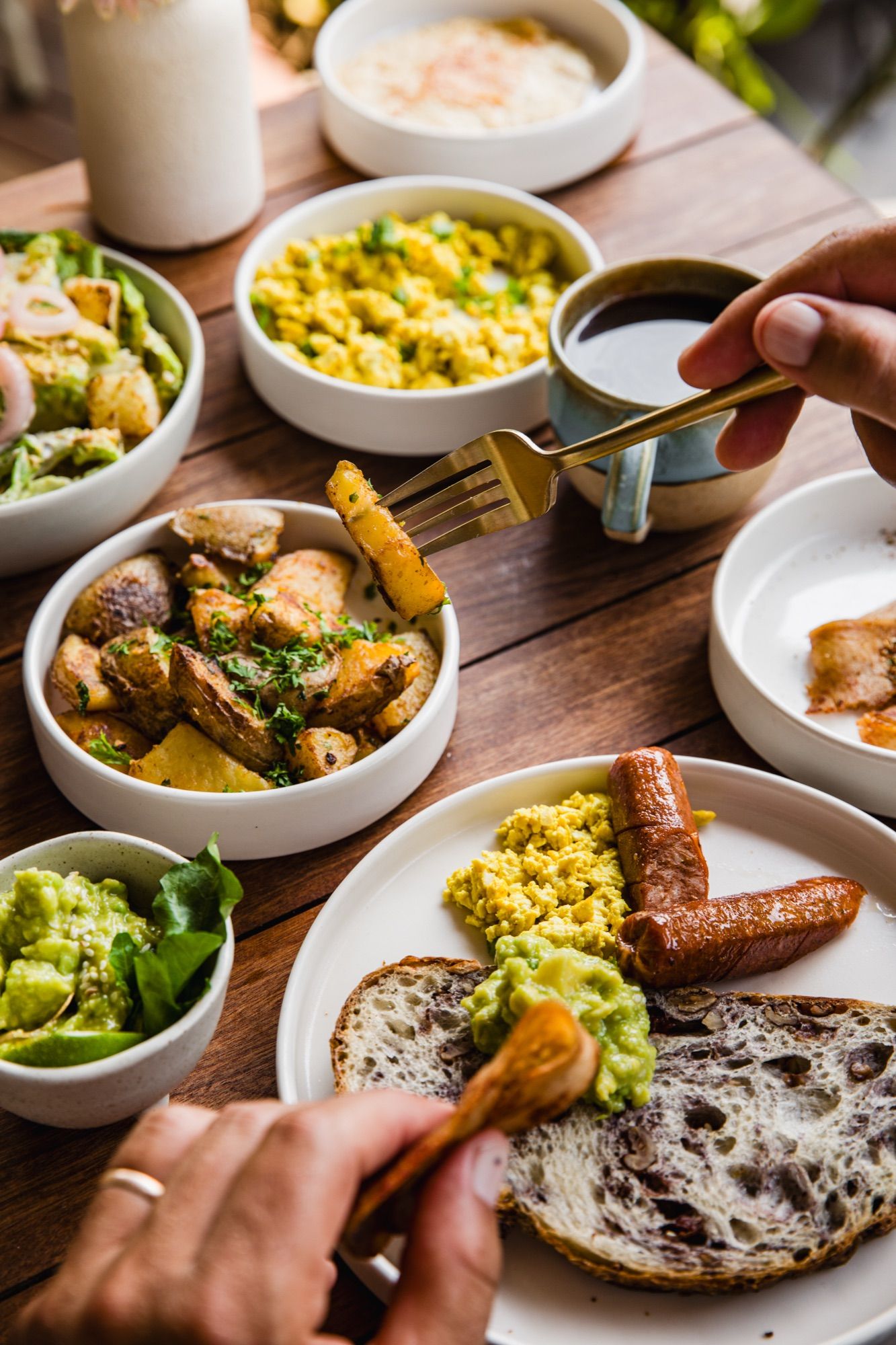 A shared breakfast table: tofu scramble, roast potatoes, avocado, sausages, sourdough and coffee being poured